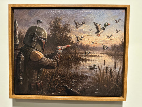 A framed painting depicting a Star Wars Mandalorian in armor aiming a blaster at flying mallards by a marsh, shown hanging on a light wall.