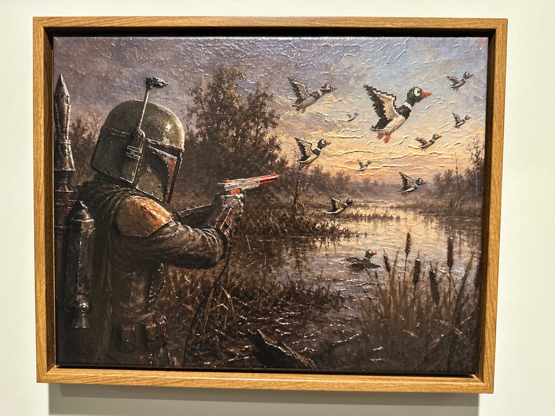 A framed painting depicting a Star Wars Mandalorian in armor aiming a blaster at flying mallards by a marsh, shown hanging on a light wall.