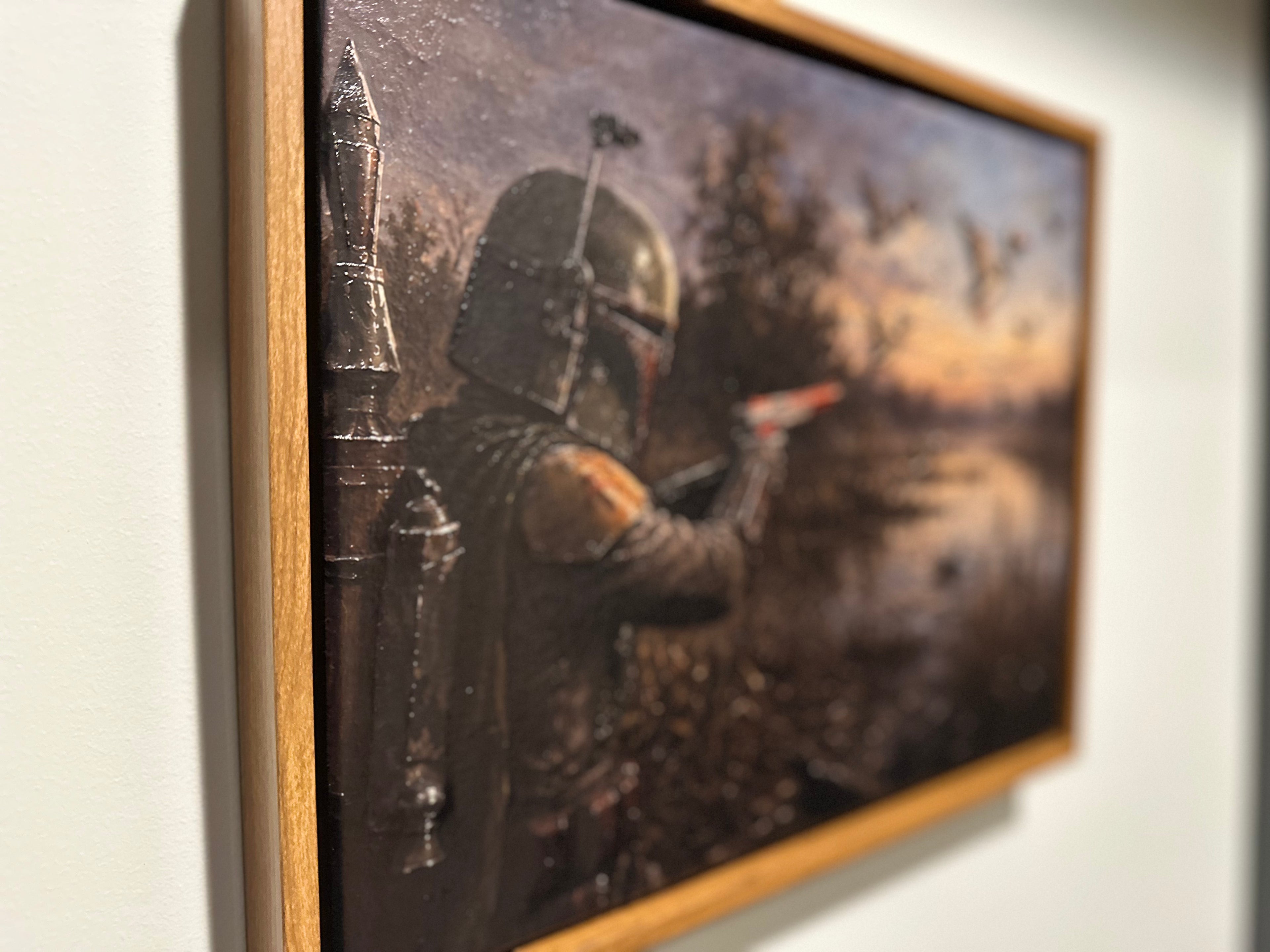 A framed painting depicting a Star Wars Mandalorian in armor aiming a blaster at flying mallards by a marsh, shown hanging on a light wall.
